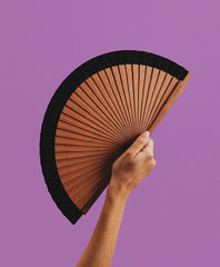 A hand holding the open Wood Black hand fan against a purple background