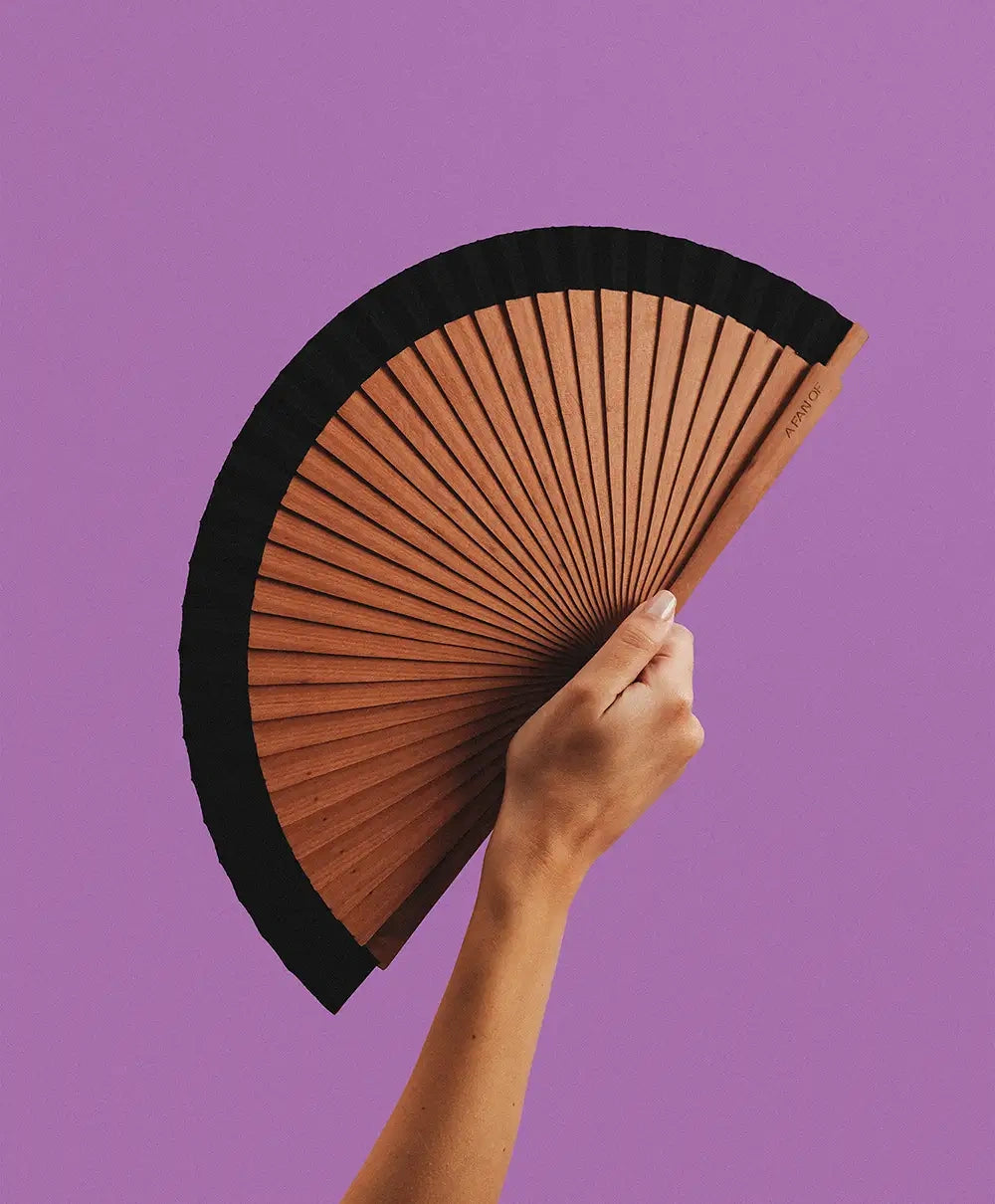 A hand holding the open Wood Black hand fan against a purple background