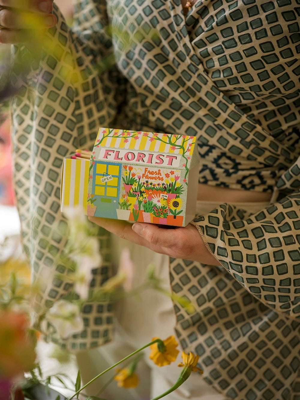 A hand holding the Florist house matchbox beside flowers